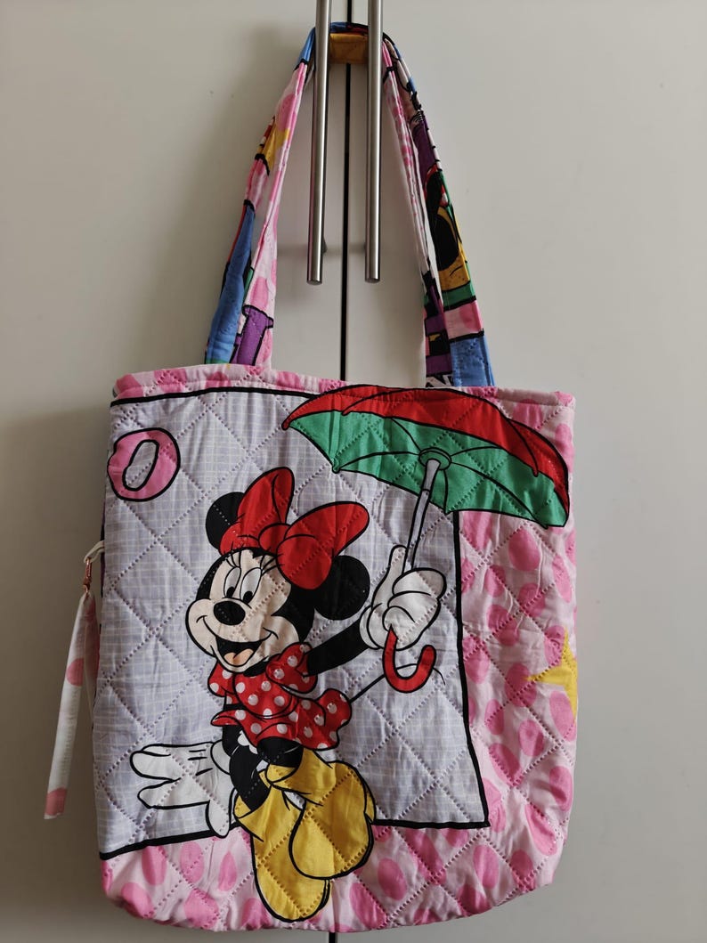 Może przedstawiać: Pikowana torba na zakupy z Minnie Mouse trzymającą parasol. Torba ma białe tło z rombowym wzorem i r&oacute;żowo-białą obw&oacute;dką w kropki. Uchwyty są wielokolorowe.