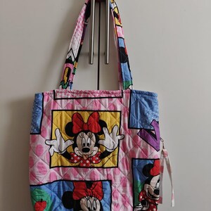 Może przedstawiać: Pikowana torba typu tote z Myszką Minnie w r&oacute;żnych pozach. Torba ma r&oacute;żowe tło z rombowym wzorem i kolorowymi kwadratami. Uchwyty i wykończenie są wzorzyste z postaciami z kresk&oacute;wek. Do boku przymocowany jest uchwyt zamka błyskawicznego.
