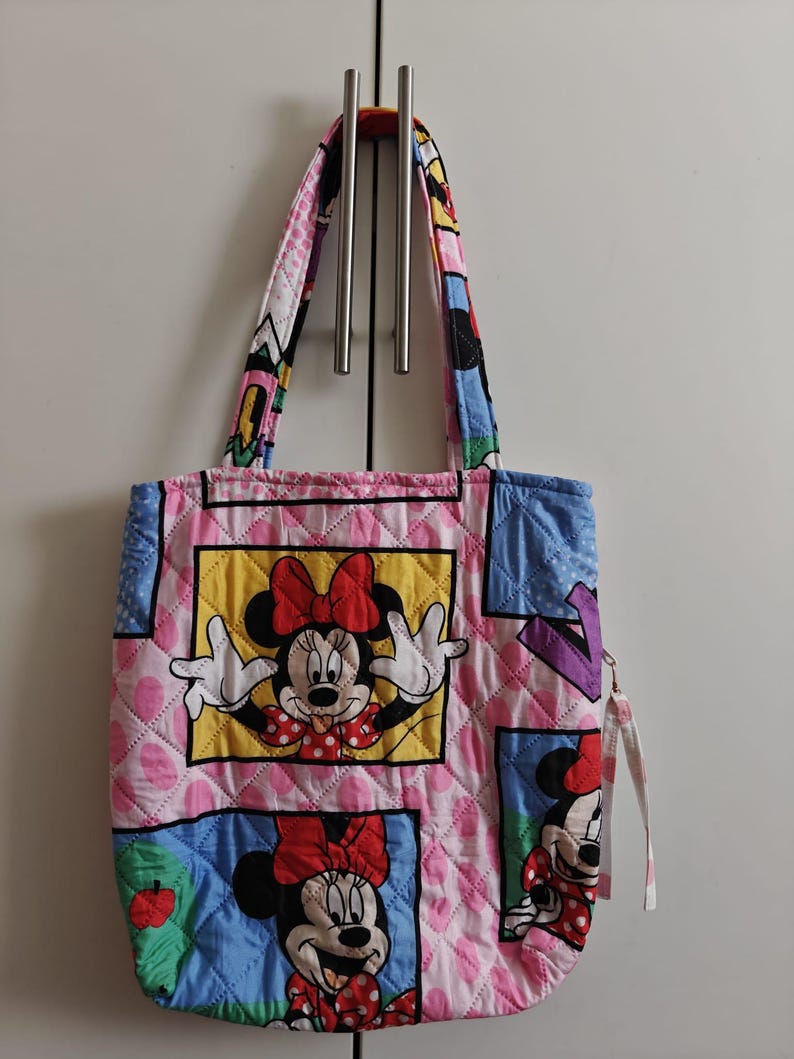 Może przedstawiać: Pikowana torba typu tote z Myszką Minnie w r&oacute;żnych pozach. Torba ma r&oacute;żowe tło z rombowym wzorem i kolorowymi kwadratami. Uchwyty i wykończenie są wzorzyste z postaciami z kresk&oacute;wek. Do boku przymocowany jest uchwyt zamka błyskawicznego.