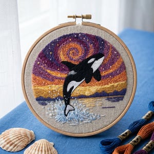 May include: A circular wooden embroidery hoop containing a hand-stitched scene of a black and white orca whale leaping from water. The background features a vibrant sunset with purple, orange, and yellow swirls, accented by small white beads.