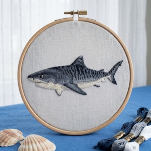 May include: A circular wooden embroidery hoop containing a light grey fabric panel with a detailed hand-stitched tiger shark. The shark features dark grey and black stripes on its back with a white underbelly. Two seashells and three embroidery floss skeins.