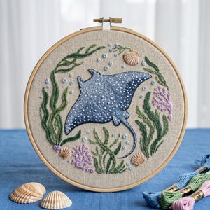 May include: A circular wooden embroidery hoop containing a fabric panel with a hand-stitched design of a blue spotted eagle ray surrounded by green seaweed, light purple coral, and small tan seashells, accented with light blue embroidered bubbles.