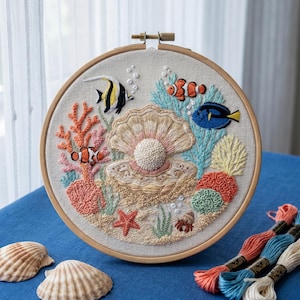 May include: A circular wooden embroidery hoop containing a fabric panel with a hand-stitched underwater scene featuring a large open seashell with a central pearl, surrounded by colorful coral, a starfish, and various tropical fish species.