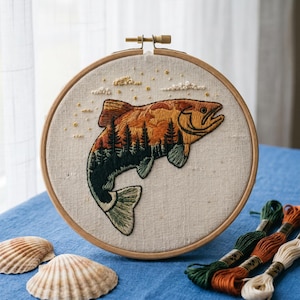 Puede incluir: Un bastidor de bordado circular de madera que contiene un panel de tela con un diseño cosido a mano de un pez saltando. El cuerpo del pez presenta un paisaje de pinos verde oscuro contra un cielo al atardecer naranja. Nubes y estrellas están bordadas arriba.