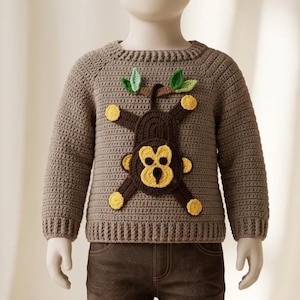 Peut inclure: Un pull en crochet marron clair à manches longues orné d'une applique de singe marron foncé suspendu à une branche marron avec deux feuilles vertes. Le singe a un visage, des mains et des pieds jaunes. Le pull présente un col et des poignets côtelés.