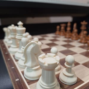 Könnte beinhalten: Nahaufnahme eines Schachspiels mit weißen und braunen Figuren auf einem Holzbrett. Die weißen Figuren, darunter ein Turm, ein Springer und ein Bauer, stehen im Vordergrund. Das Schachbrett hat braune und beige Quadrate.