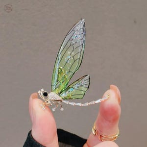 Op de afbeelding: Een libellebroche met een goudkleurig lichaam en transparante, groen getinte vleugels. De vleugels hebben een delicaat, geaderd patroon en zijn versierd met iriserende glitter. De broche heeft een zwart oog en heldere kristallen accenten.