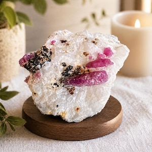 Può includere: Un campione di quarzo bianco grezzo con vibranti cristalli di rubino rosa. Il minerale è posto su un piccolo supporto rotondo di legno. Macchie scure e piccole macchie marroni sono visibili sul quarzo. Una candela accesa e una pianta in vaso sono sullo sfondo.