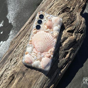 May include: A clear smartphone case decorated with an assortment of natural seashells in shades of cream, tan, and soft pink, interspersed with small white spherical pearls. The case features three circular camera lens cutouts on the upper left side.