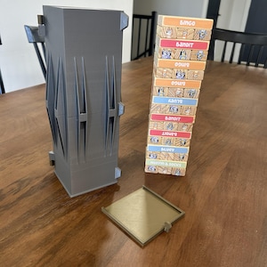 May include: A gray 3D-printed storage container with a matching tray and a tower of wooden blocks. The blocks are labeled with names and images, stacked in a leaning tower. The container has a geometric design.
