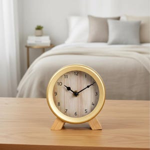 May include: A round analog clock with a gold-colored metal frame and two small wooden feet. The clock face features a light wood grain pattern with black numerals from 1 to 12 and two black hands indicating the time in a circular design.
