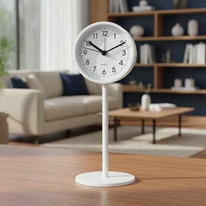 May include: A white analogue table clock featuring a circular face with black numerals from 1 to 12, black hour and minute hands, and the word QUARTZ printed in black. The clock is mounted on a slender white vertical post and a round white base.