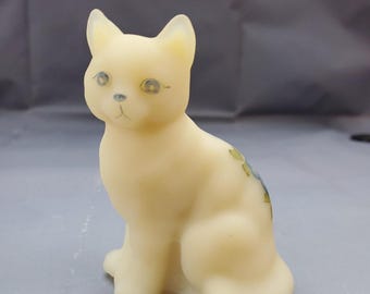 Puede incluir: Una pequeña figura de gato sentada hecha de vidrio esmerilado translúcido de color amarillo pálido. El gato tiene ojos azules pintados, una nariz azul pequeña y un delicado patrón floral verde y azul pintado en su espalda. La superficie es lisa y mate.