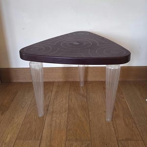 May include: A triangular side table with a dark brown top featuring a circular pattern. The table has three clear, fluted legs. The table is approximately 20 inches wide and 18 inches tall.