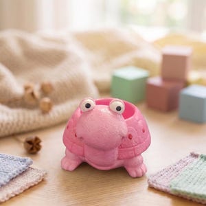 May include: A pink turtle-shaped planter with large, googly eyes. The planter is made of a textured material and has a round opening for plants. The turtle has four small feet and a detailed shell. The background includes wooden blocks and a soft blanket.