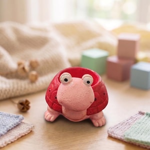 Könnte beinhalten: Ein roter, schildkrötenförmiger Pflanztopf mit rosa Gesicht und Füßen. Der Pflanztopf hat große, wackelige Augen und eine runde Öffnung für Pflanzen. Der Schildkröten-Pflanztopf ist aus Keramik und steht auf einem Holztisch.