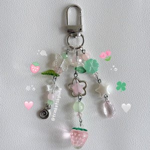 May include: A silver-toned metal keychain featuring three dangling strands of beads. The charms include a translucent butterfly, a pink strawberry, a flower-shaped metal charm, a white star, and various green and pink spherical beads.
