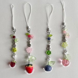 May include: Four phone charms featuring white cord loops and silver-tone metal hardware. Each charm includes a variety of beads, including green leaves, small flowers, and fruit-shaped pendants: a red raspberry, a strawberry, a blueberry, and a peach.