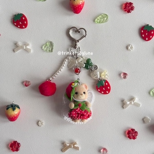 May include: A keychain featuring a small bear figurine wearing a red strawberry-patterned dress and a green leaf hat. The charm is attached to a silver heart-shaped clasp with dangling white pearls, a red pom-pom, and strawberry-shaped beads.