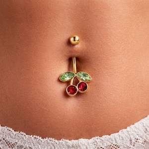 Puede incluir: Anillo para el ombligo dorado con diseño de cereza. La cereza tiene dos gemas rojas y dos hojas de gemas verdes. La bola superior también es dorada. La joya se lleva en el abdomen de una persona.