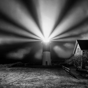Puede incluir: Fotografía en blanco y negro de un faro por la noche, con un haz de luz brillante que irradia hacia afuera. Una casa y una valla son visibles en el fondo. La imagen está tomada al anochecer o por la noche.