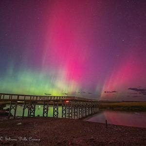 Może przedstawiać: Nocny krajobraz z drewnianym mostem nad wodą, pod zorzą polarną. Niebo prezentuje gradient fioletu, różu i zieleni, z widocznymi gwiazdami. Most widoczny jako sylwetka.