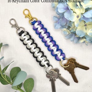 May include: Two paracord keychains with metal clasps and attached keys. One keychain features a black, gray, and white woven pattern with a silver-tone clasp. The other features a blue, purple, and white pattern with a gold-tone clasp. Text reads: 16 Keychain Color Combinations Available.