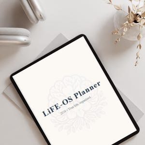 May include: A digital tablet displaying the text LiFE-OS Planner 2026 Your life, organized. The screen features a faint brain-shaped floral graphic. The brand name BRAINSBLOOM is visible at the top, accompanied by silver headphones and a small floral vase.