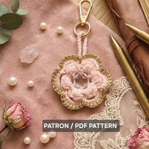Peut inclure: Un porte-clés en forme de fleur rose crochetée, avec une bordure et un fermoir dorés. La fleur a un centre perlé transparent. L'image comprend également des perles, des roses séchées et le texte "PATRON / PDF PATTERN".