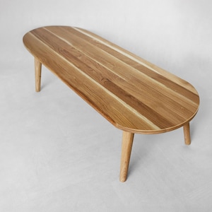 Peut inclure: Table basse ovale en bois clair, avec quatre pieds effilés. Le plateau est composé de planches de bois disposées dans le sens de la longueur. La table présente un veinage naturel et se détache sur un fond blanc.