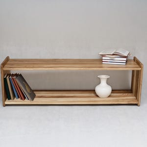 Peut inclure: Une étagère basse rectangulaire en bois à deux niveaux horizontaux. Le meuble présente une finition en grain de bois naturel brun clair. Sur l'étagère supérieure repose une pile de livres. L'étagère inférieure contient des livres et un vase blanc.
