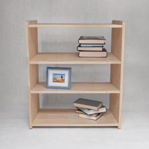 May include: A light-colored wooden bookshelf with four open shelves. The top shelf holds a stack of three books. The second shelf contains a small rectangular picture frame with a blue border. The bottom shelf holds a stack of three books.