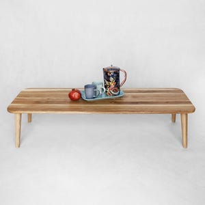 Peut inclure: Une table basse rectangulaire en bois avec des coins arrondis et quatre pieds. Sur la table, un plateau bleu avec deux tasses bleues, une cruche décorative à motifs floraux et une grenade rouge.