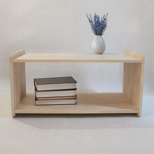 May include: A light-coloured wooden shelf with two horizontal tiers. A white ribbed vase containing purple lavender flowers sits on the top shelf, while a stack of three books with dark covers rests on the bottom shelf of the wooden unit.