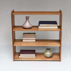 Op de afbeelding: Een houten boekenplank met vier niveaus en een natuurlijke bruine afwerking. De planken bevatten twee stapels boeken met donkere kaften en lichte pagina's, een lichtroze keramische vaas bovenop en een saliegroene keramische vaas onderaan.