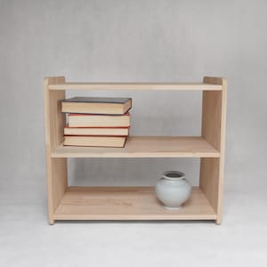 Puede incluir: Una estantería de madera de color claro con dos estantes abiertos. El estante superior sostiene una pila de cuatro libros con cubiertas oscuras y páginas claras. El estante inferior contiene un pequeño jarrón de cerámica gris claro, redondo y de cuello estrecho.