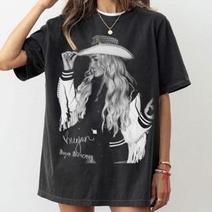 May include: Black graphic t-shirt with a black and white image of a person in a cowboy hat and jacket. The text "Megan" and "Megan Moroney" are written in cursive. The shirt has short sleeves and a vintage look.