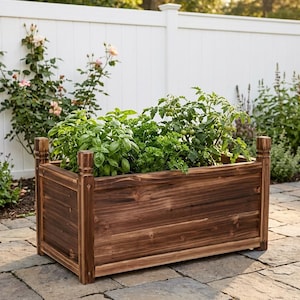 Puede incluir: Una jardinera rectangular de madera de color marrón oscuro con postes de esquina verticales. La jardinera presenta una textura de veta de madera visible y contiene plantas verdes de hoja, incluyendo albahaca y plantas de tomate con frutos.