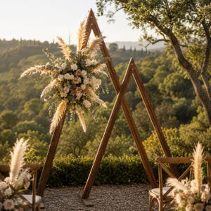 May include: A large wooden triangular wedding arch frame featuring a floral arrangement of white roses, eucalyptus leaves, and tall pampas grass plumes. The structure consists of two connected triangular frames made of dark brown stained wood.