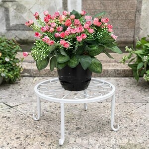 May include: A black plastic pot filled with artificial pink rose buds and green leaves, resting on a white metal plant stand. The stand features a circular top with a decorative scrollwork pattern and four curved legs that extend to the ground.