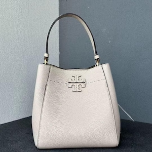 Könnte beinhalten: Eine hellbeige Handtasche mit einem oberen Griff und einem Logo auf der Vorderseite. Die Tasche hat eine strukturierte Form und eine Naht am oberen Rand. Der Griff ist schwarz mit goldfarbenen Details.