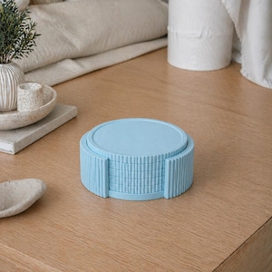 May include: A stack of light blue coasters with a ribbed edge design. The coasters are stacked neatly on a wooden table. The set includes a top coaster and several individual coasters, perfect for protecting surfaces from drink rings.
