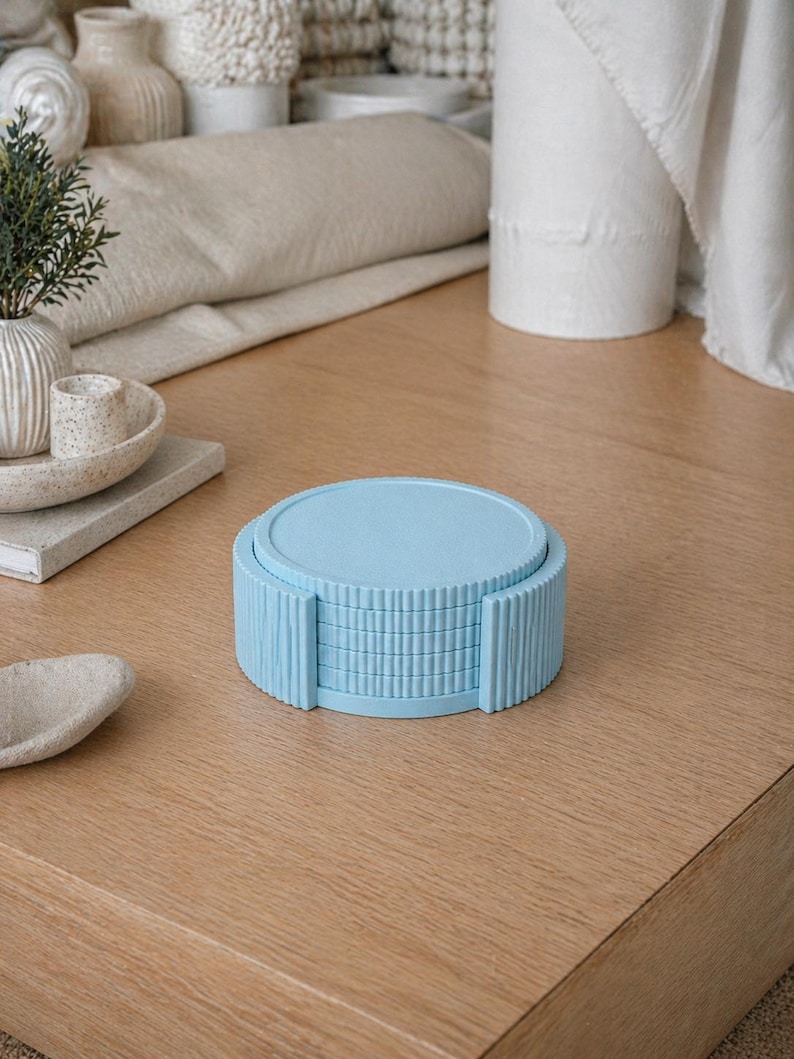 May include: A stack of light blue coasters with a ribbed edge design. The coasters are stacked neatly on a wooden table. The set includes a top coaster and several individual coasters, perfect for protecting surfaces from drink rings.