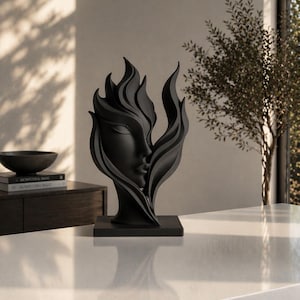 May include: A black sculpture of a woman's face intertwined with stylized flames. The artwork is set on a square base and placed on a white countertop. The sculpture is a decorative piece for home decor.
