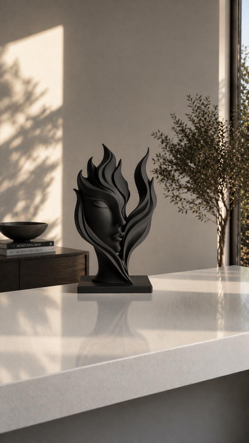 May include: A black sculpture of a woman's face intertwined with stylized flames. The artwork is set on a square base and placed on a white countertop. The sculpture is a decorative piece for home decor.