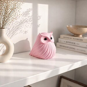 May include: A pink owl figurine with black eyes sits on a white shelf. The owl has a textured, feathered design. A vase with dried flowers and books are also on the shelf. The books are titled "Aesthetic Interiors" and "Minimalist Living".