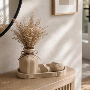May include: A beige ceramic vase with dried flowers, a small heart-shaped object, and a candle holder on a matching tray. The vase has a brown ribbon tied around its neck. The items are on a wooden table.