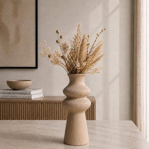 May include: A beige, ribbed ceramic vase with a bulbous shape, holding a bouquet of dried beige and tan flowers. The vase is on a light-colored surface, with a wooden cabinet and artwork in the background.