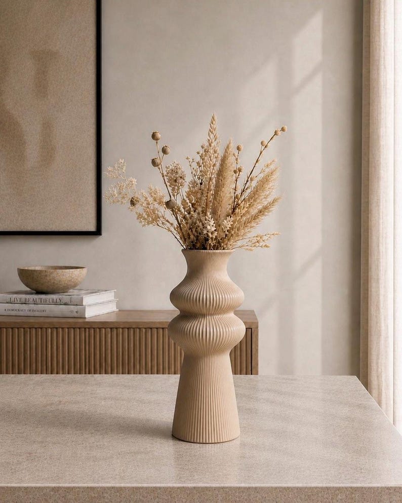 May include: A beige, ribbed ceramic vase with a bulbous shape, holding a bouquet of dried beige and tan flowers. The vase is on a light-colored surface, with a wooden cabinet and artwork in the background.