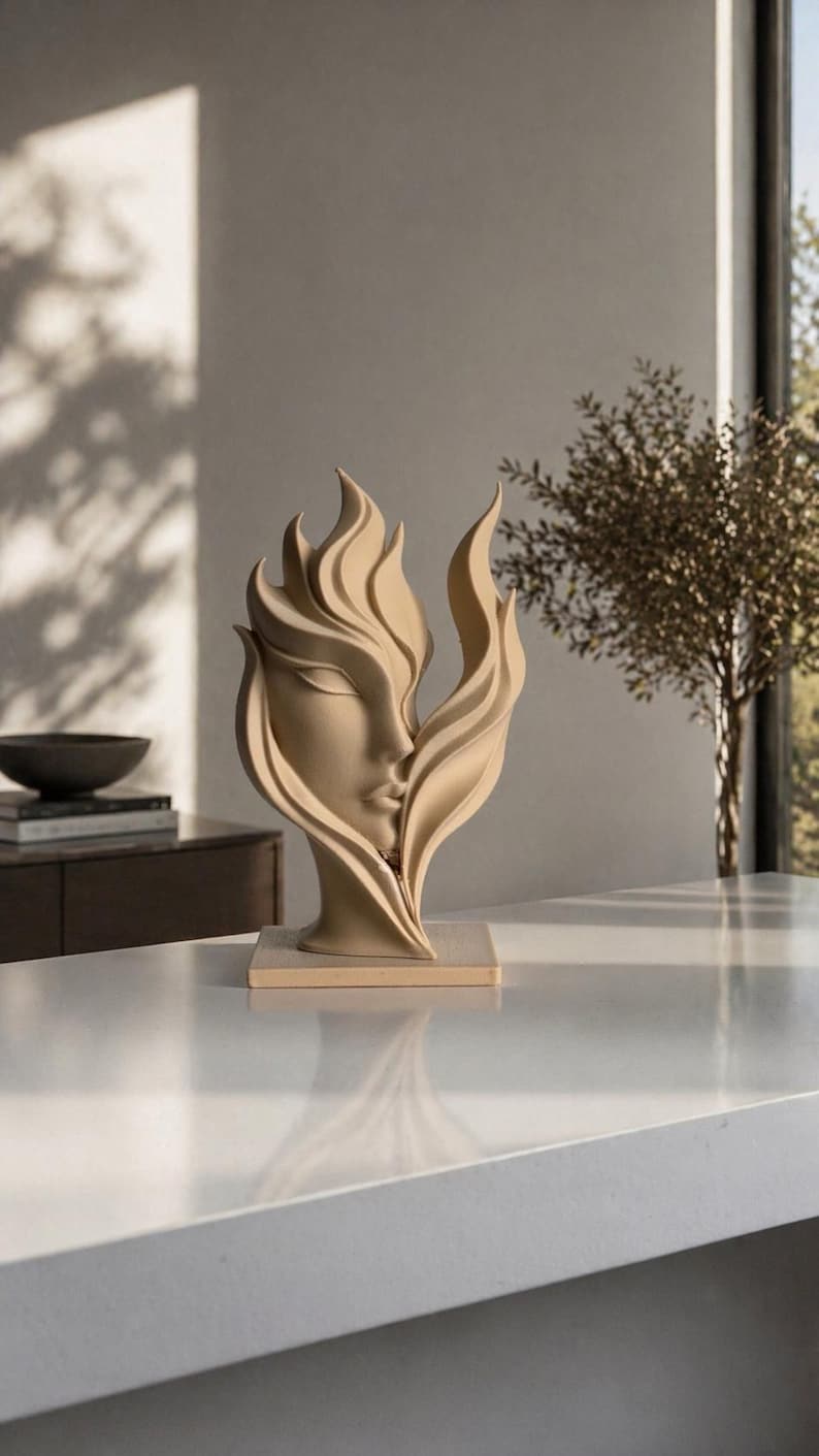 May include: A beige sculpture of a woman's face with flowing, flame-like hair. The artwork is set on a light-colored rectangular base. The sculpture is displayed on a white countertop, with a blurred background featuring a window and a small tree.